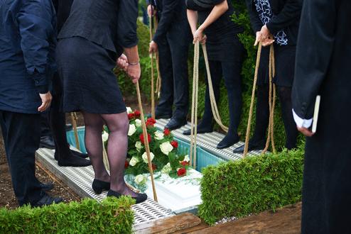 Teilnehmer einer Trauerfeier halten Seile, um Blumenschmuck auf einen Sarg in einer Grabstelle abzusenken.